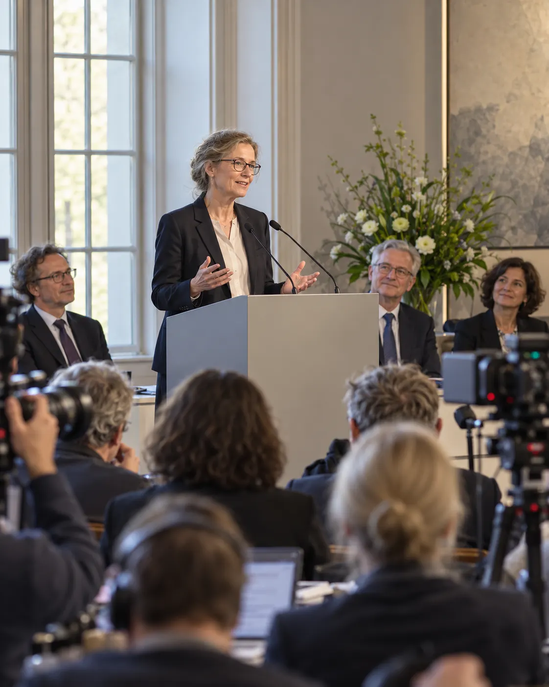AI-generated generic reconstruction of a prize announcement press conference, not a real Nobel event or identified people. Generated 19 October 2026. Prompt: “Photorealistic vertical 4:5 editorial reconstruction of a Nobel Prize announcement press conference: a scientist speaking at a plain lectern in a modern European institute press room, journalists listening, microphones and cameras, respectful candid scene. Generic fictional people, not a real Nobel event. No logos, no readable text, no medals.”