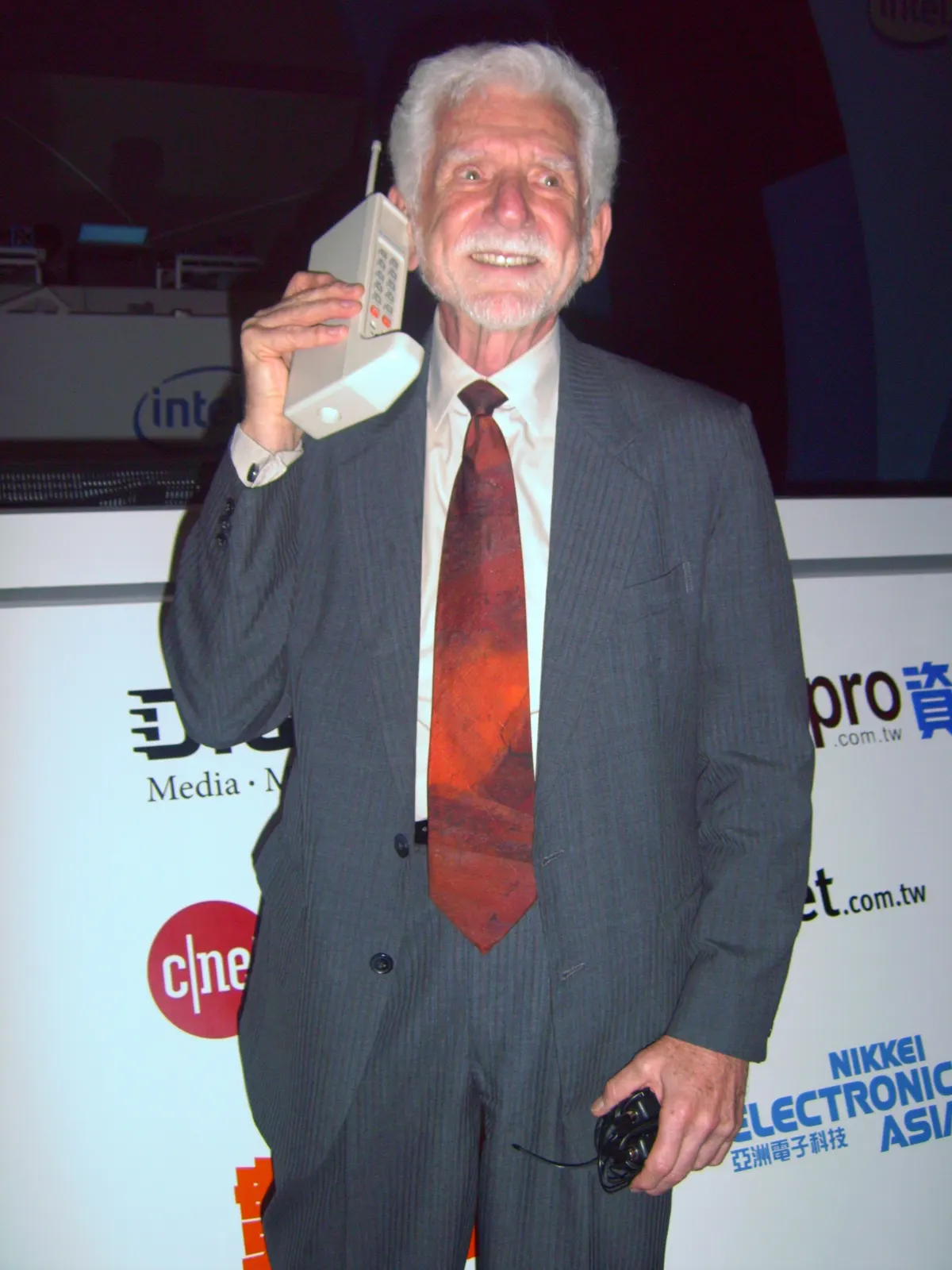 documentary photograph of Martin Cooper with a DynaTAC prototype at e21 Forum in Taipei, 5 June 2007. Photographer: Rico Shen. Original page: https://commons.wikimedia.org/wiki/File:2007Computex_e21Forum-MartinCooper.jpg. CC BY-SA 3.0 (or CC BY-SA 2.5 Taiwan/GFDL as offered on the page); attribution: “Rico Shen / Wikimedia Commons / CC BY-SA 3.0”; no edits.