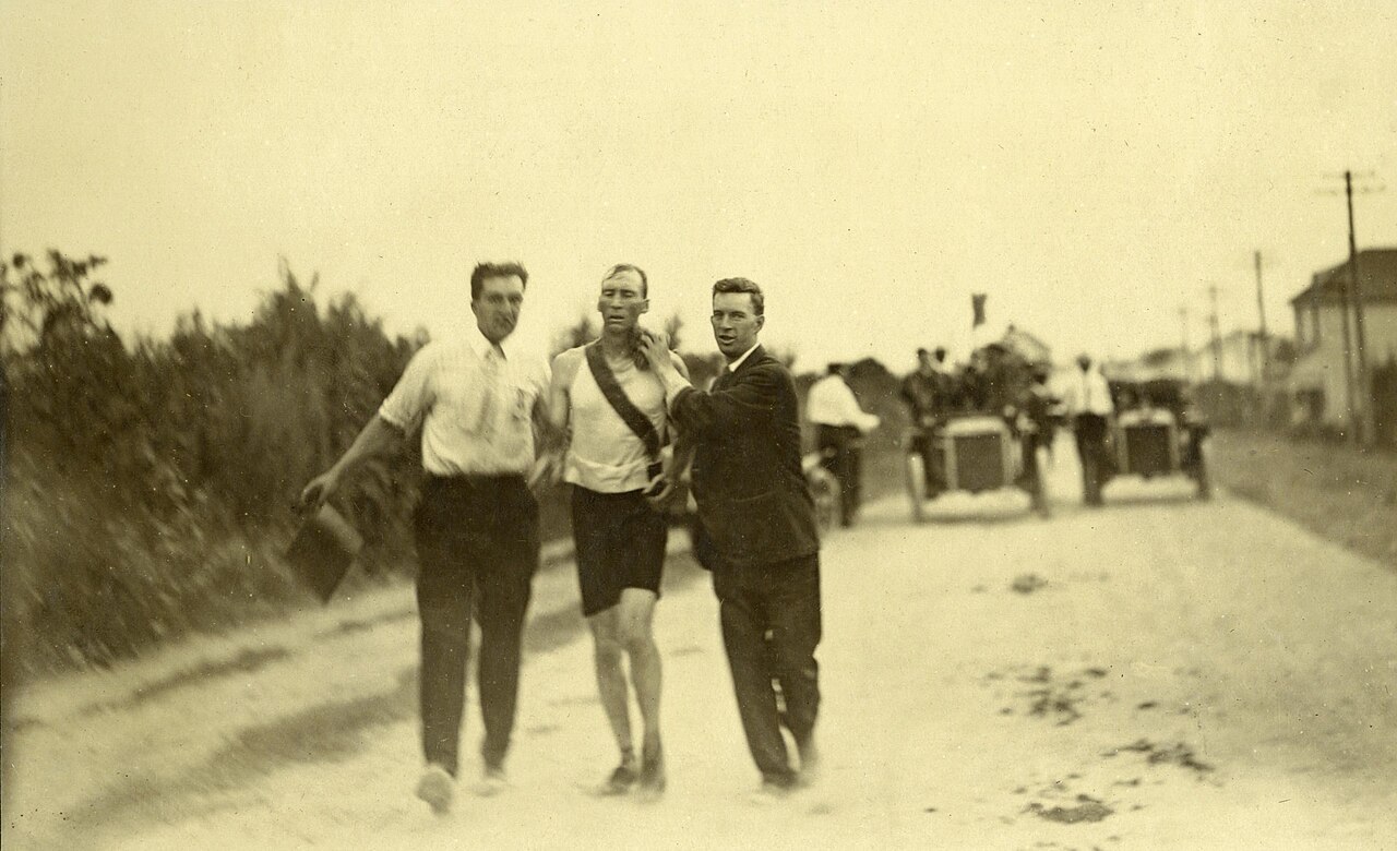 Documentary photo of Hicks being supported during the race, attributed to unknown photographer; Missouri History Museum collection via Wikimedia Commons, public domain in the United States. https://commons.wikimedia.org/wiki/File:Marathon_Hicks1904.jpg