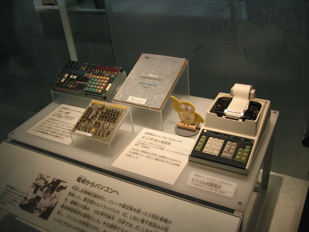 Museum display showing a Busicom calculator and a 4004-based board, photographed in 2007 by Ruud Dingemans; the photographer released the image worldwide into the public domain. No edits. https://commons.wikimedia.org/wiki/File:Busicom.JPG
