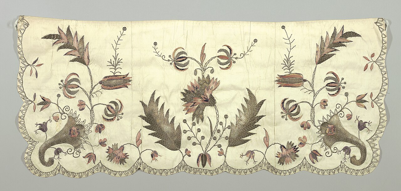 Short wide apron of white embroidered in silver thread, rose, violet and white. Design is a large central flower spray of carnation and lily with leaves in silver thread. Corner design of cornucopia with sprays of tulips and lilies. Bottom and sides have a subtle scallops bordered in a sawtooth pattern in silver thread.. Original file: [Apron (France), early 18th century (CH 18386997).jpg](https://commons.wikimedia.org/wiki/File%3AApron_%28France%29%2C_early_18th_century_%28CH_18386997%29.jpg). Creator: Unknown artist Unknown artist. License: Public domain (https://commons.wikimedia.org/wiki/File%3AApron%20%28France%29%2C%20early%2018th%20century%20%28CH%2018386997%29.jpg). Attribution: Unknown artist Unknown artist. Downloaded from the verified Commons file page; unaltered image. Checked 2026-10-25.