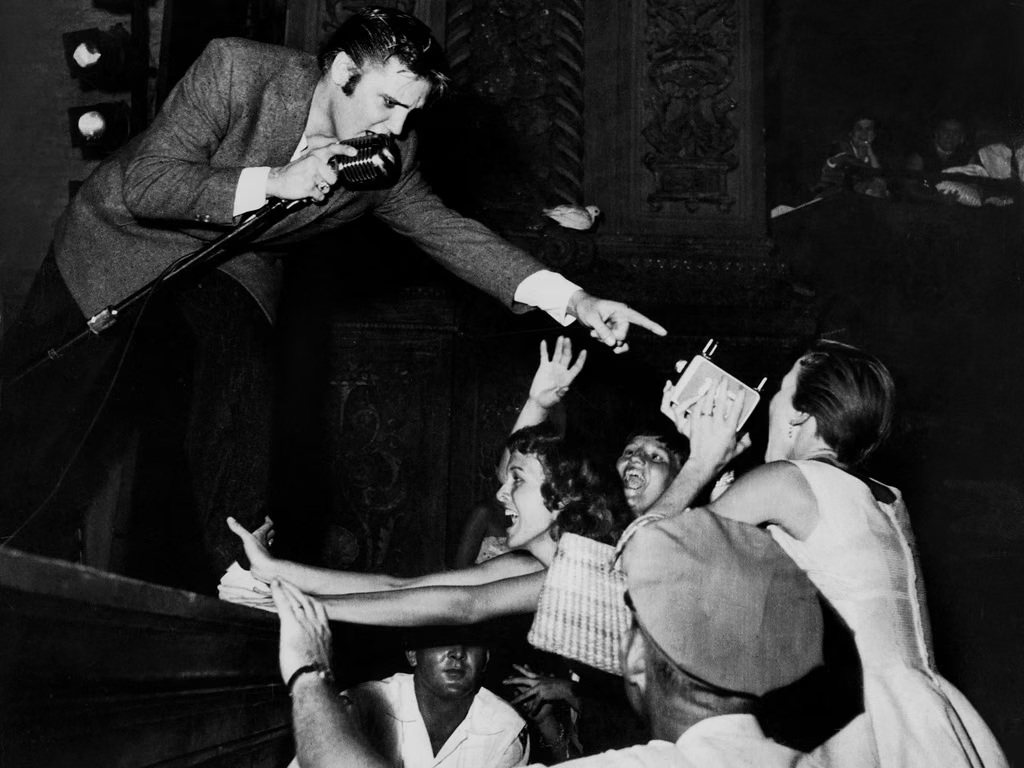 Presley performing at Miami’s Olympia Theater, Aug. 3, 1956. Don Wright for Miami Herald; Wikimedia Commons file page https://commons.wikimedia.org/wiki/File:Elvis_Presley_at_Olympia_Theater,_Aug_3,_1956.jpg; U.S. public domain, copyright not renewed per file page. No attribution required; credit Don Wright/Miami Herald when practical.